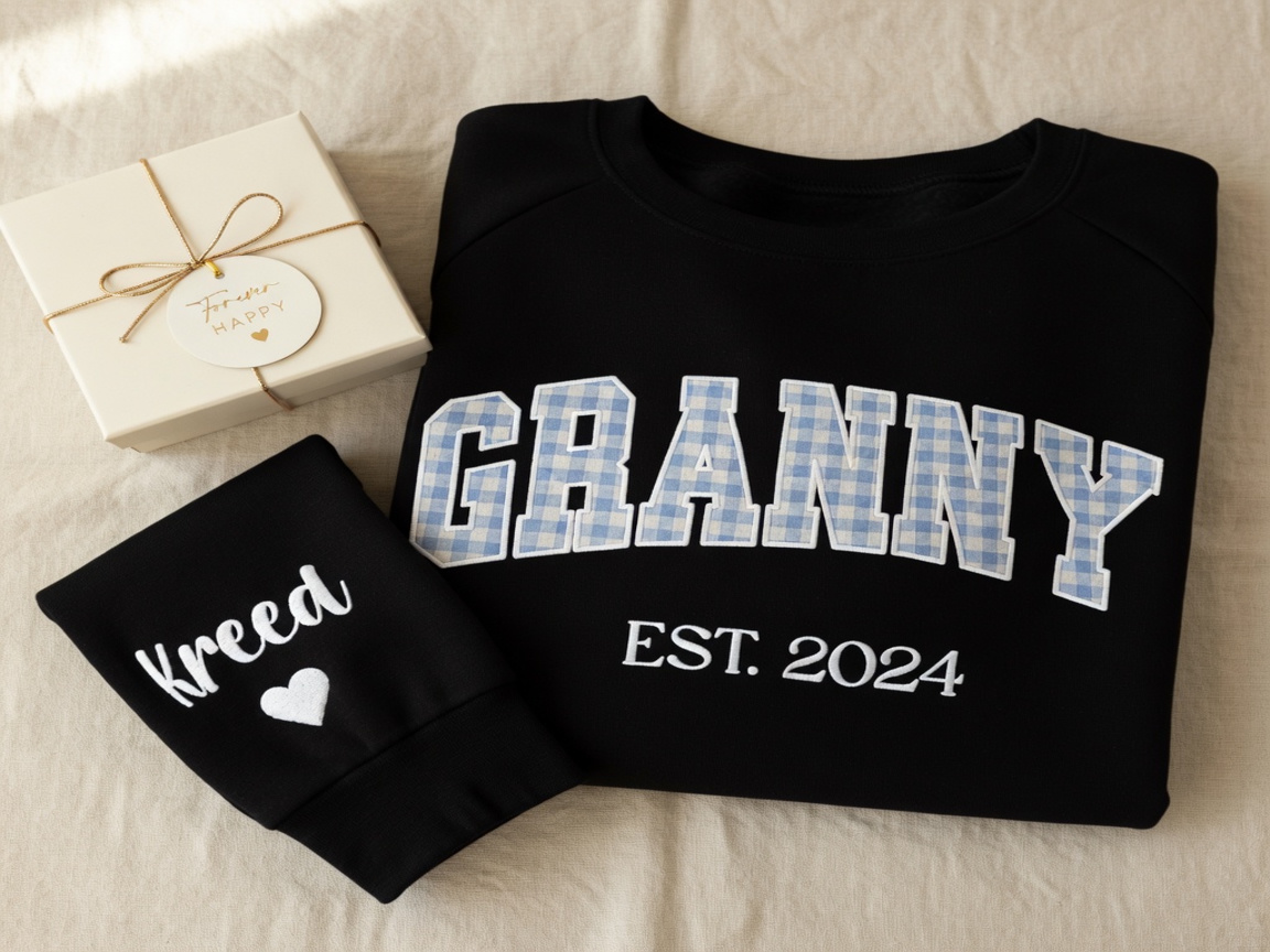 Trina's handmade GRANNY EST. 2024 sweatshirt — gingham letters, the custom name Kreed lettered on the sleeve with a small heart — styled flat lay with gift box